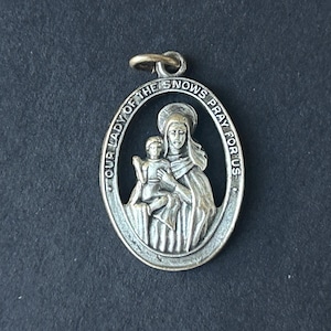 May include: Oval silver pendant featuring an embossed image of the Virgin Mary holding a child, with the text "OUR LADY OF THE SNOWS PRAY FOR US" around the edge. The pendant has a small loop at the top for a chain.