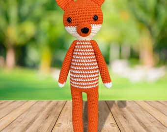 May include: A handmade orange and white crochet fox doll. The fox has a white face and chest, with black eyes and a striped torso. The fox is standing on a wooden surface with a blurred green background.