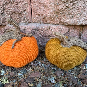May include: Two crocheted pumpkins, one orange and one mustard yellow, with brown stems, sitting on a bed of brown leaves and rocks.