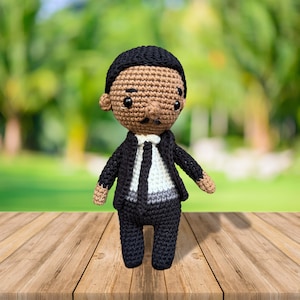 Peut inclure: Une poupée en crochet avec une peau brune, des cheveux noirs et un costume noir. La poupée porte une chemise blanche et une cravate noire. La poupée est debout sur une surface en bois.