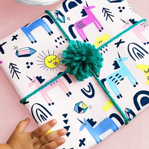 May include: A white gift wrap with a pink background and a repeating pattern of unicorns, rainbows, stars, and geometric shapes. The gift is tied with a green ribbon and a green pom-pom.