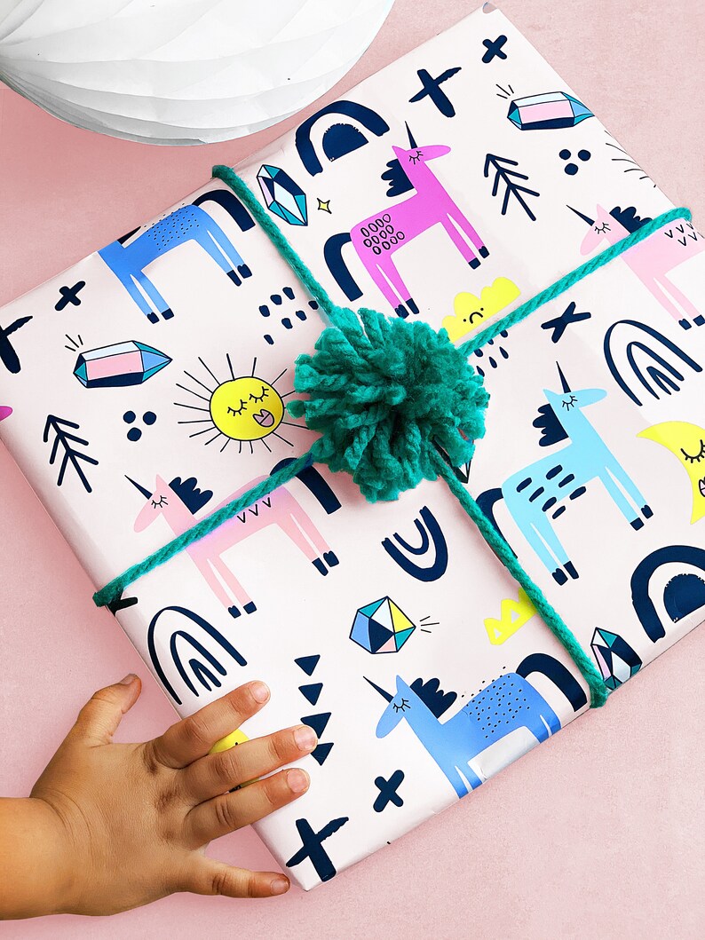 May include: A white gift wrap with a pink background and a repeating pattern of unicorns, rainbows, stars, and geometric shapes. The gift is tied with a green ribbon and a green pom-pom.