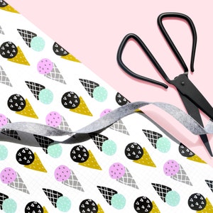 May include: White wrapping paper with a repeating pattern of ice cream cones, circles, and geometric shapes in black, gray, gold, pink, and light blue. A gray ribbon and black scissors are on top of the paper.