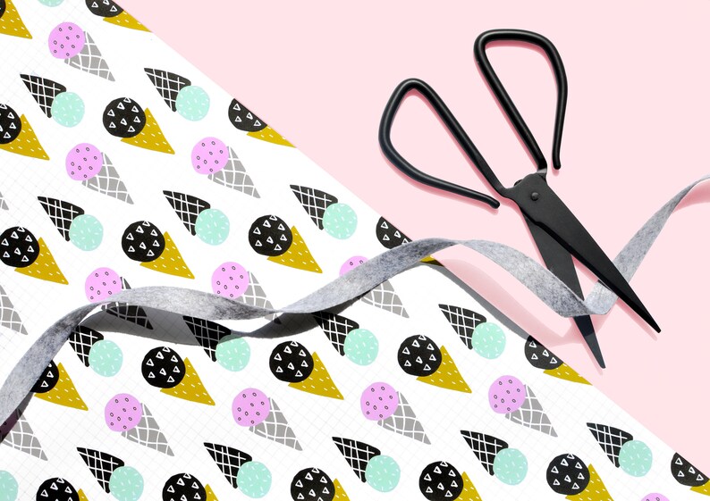 May include: White wrapping paper with a repeating pattern of ice cream cones, circles, and geometric shapes in black, gray, gold, pink, and light blue. A gray ribbon and black scissors are on top of the paper.