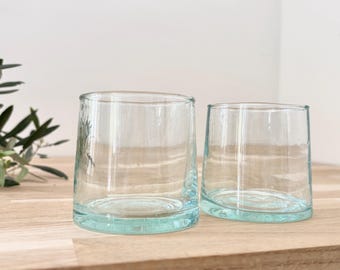 May include: Two clear, rounded drinking glasses with a slight aqua tint at the base. The glasses are made of a thick glass material and sit on a light-colored wooden surface. The glasses are empty and ready to be filled.