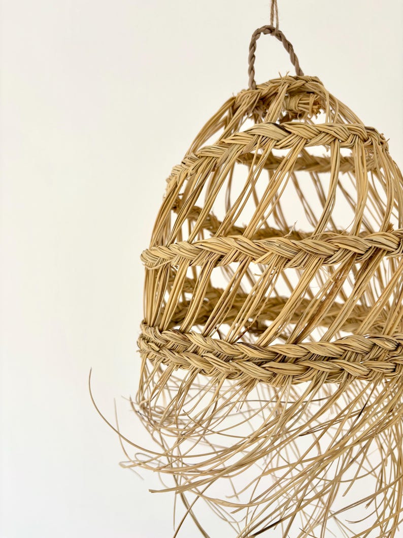 May include: A woven, natural-toned pendant light fixture. The light has a rounded, cage-like design with a braided top and a hanging loop. The bottom of the light has long, loose strands of the same material.