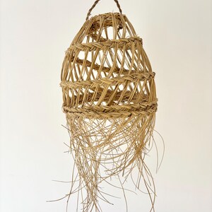 May include: A woven basket with a teardrop shape, suspended from a brown cord. The basket is made of light brown, natural fibers, with a woven pattern and long, trailing strands at the bottom. The basket has a handle at the top.