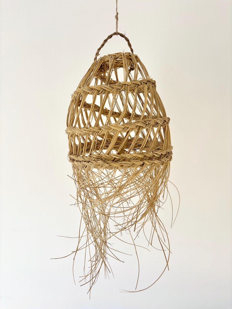 May include: A woven basket with a teardrop shape, suspended from a brown cord. The basket is made of light brown, natural fibers, with a woven pattern and long, trailing strands at the bottom. The basket has a handle at the top.