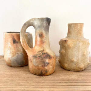 Op de afbeelding: Drie keramische vazen in verschillende vormen en maten. De vazen zijn lichtbruin van kleur met een gemarmerd, getextureerd oppervlak.