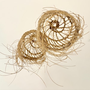 May include: Two handcrafted, spherical woven baskets made of natural-colored, dried plant fibers. The baskets have an open, airy design with spiraling and radial patterns. Strands of the fiber extend outward, creating a textured, organic aesthetic.