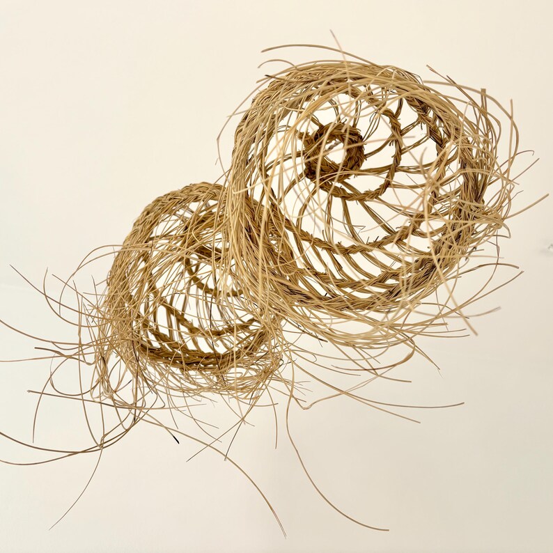 May include: Two handcrafted, spherical woven baskets made of natural-colored, dried plant fibers. The baskets have an open, airy design with spiraling and radial patterns. Strands of the fiber extend outward, creating a textured, organic aesthetic.