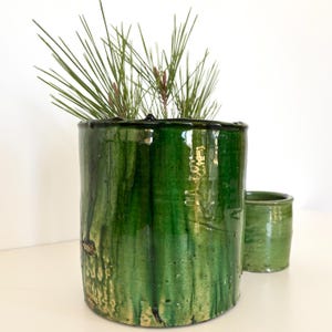 Może przedstawiać: Wysoki, cylindryczny zielony ceramiczny doniczka z błyszczącym wykończeniem, w towarzystwie mniejszego, pasującego garnka. Doniczka jest wypełniona długimi, cienkimi zielonymi igłami sosny. Garnki mają rustykalny, ręcznie robiony wygląd.
