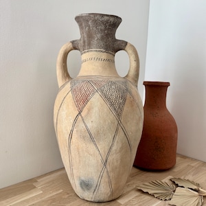 Könnte beinhalten: Eine große, zweigriffige Keramik-Amphore mit strukturierter, hellbeiger Oberfläche. Der Körper ist mit handgemalten schwarzen geometrischen Rautenmustern verziert und hat einen dunkelbraunen Hals. Dahinter steht eine kleinere, terrakottafarbene Vase.