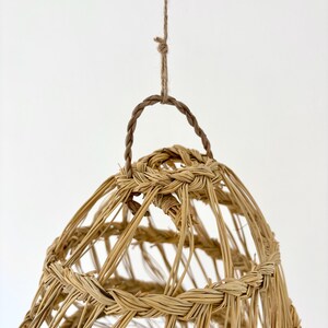 May include: A woven, bell-shaped pendant light fixture. The light fixture is made of natural-colored woven material, with a braided handle and hanging cord. The design is open, allowing light to filter through. The fixture is suspended against a white background.