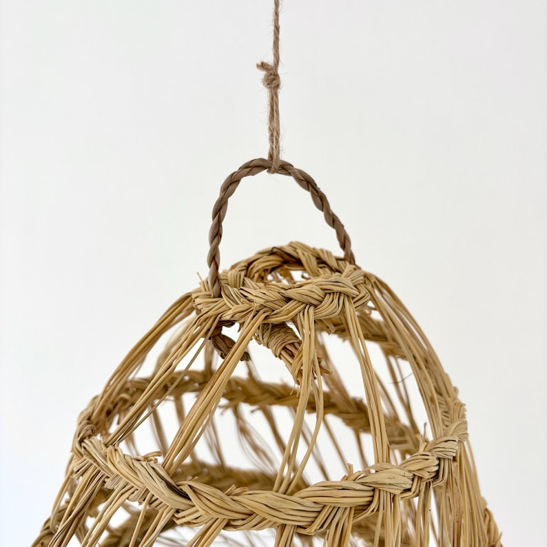 May include: A woven, bell-shaped pendant light fixture. The light fixture is made of natural-colored woven material, with a braided handle and hanging cord. The design is open, allowing light to filter through. The fixture is suspended against a white background.
