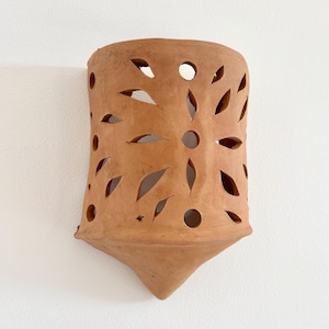 Pode incluir: Um aplique de parede de terracota com um corpo semicilíndrico e uma base pontiaguda. A superfície é decorada com uma série de formas recortadas, incluindo orifícios circulares e padrões alongados em forma de folha.