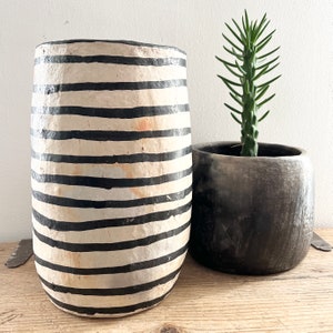 Peut inclure: Un grand vase cylindrique avec des rayures noires horizontales sur fond crème. À côté, un pot rond gris foncé contient une plante succulente verte. Les objets sont présentés sur une surface en bois, créant une esthétique rustique et naturelle.