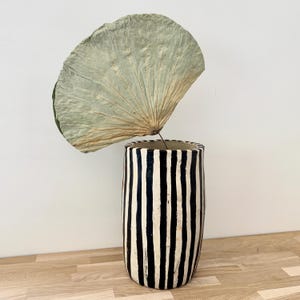 May include: A cylindrical ceramic vase featuring a hand-painted pattern of vertical black and white stripes. A single large, dried, fan-shaped lotus leaf with a pale green and tan texture is positioned inside the vase, extending upwards.