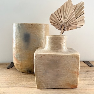 Peut inclure: Deux vases en céramique, l'un grand et rond avec un glacis brun clair, jaune et bleu, et l'autre un vase carré plus court avec un glacis beige clair. Une feuille de palmier séchée est dans le vase carré.