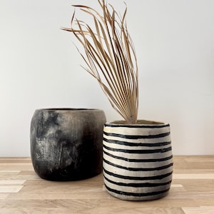 Op de afbeelding: Twee cilindervormige keramische plantenpotten. De ene pot heeft een effen, gevlekte antraciet en zwarte afwerking. De tweede pot heeft een witte basis met horizontale zwarte strepen en bevat een gedroogd, waaiervormig palmblad.