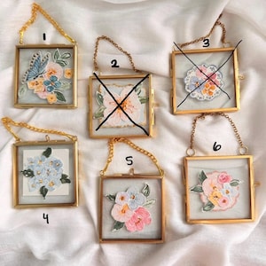 May include: Five square glass frames with gold metal edges and gold chains for hanging. Each frame contains a unique embroidered floral patch. Frame 1 features a blue butterfly and flowers, 4 shows blue blossoms, 5 displays pink and white flowers, and 6 holds a cluster of pink and white roses.