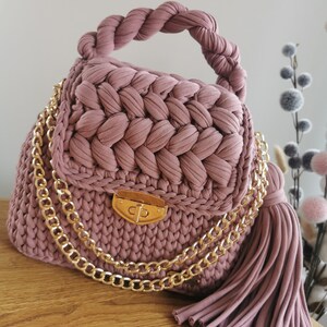 May include: A pink crocheted handbag with a gold chain strap and a tassel. The bag has a gold clasp closure.