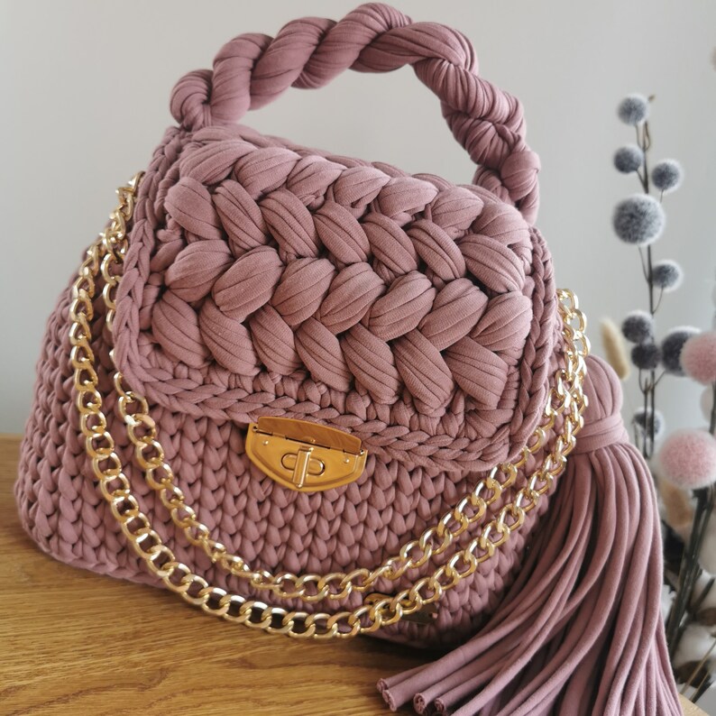 May include: A pink crocheted handbag with a gold chain strap and a tassel. The bag has a gold clasp closure.