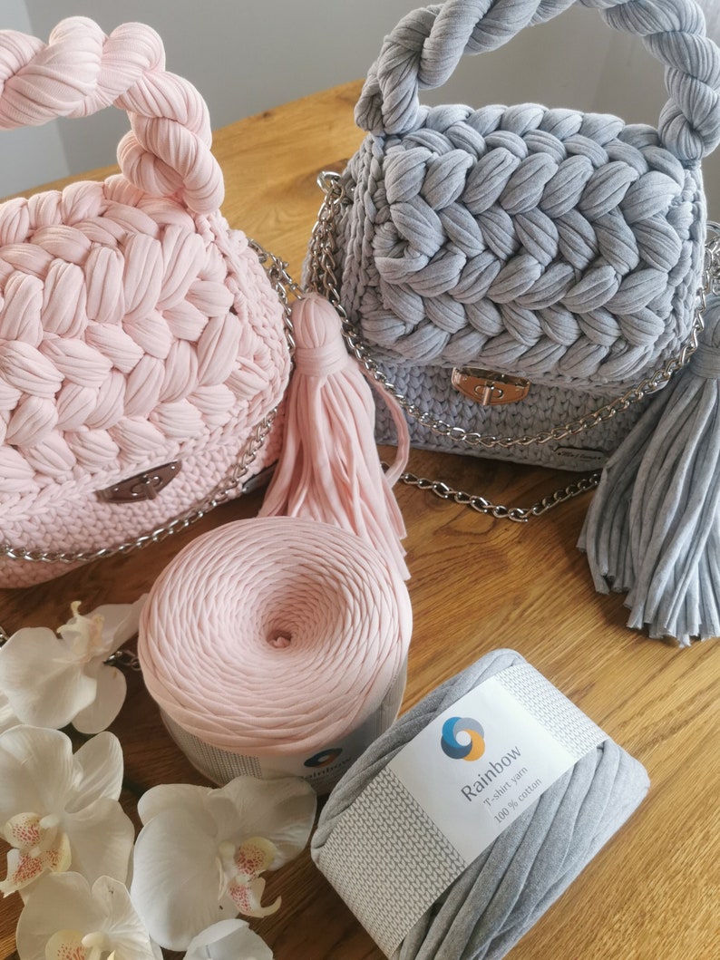 May include: Two crocheted handbags, one pink and one gray, with matching yarn skeins. The yarn is labeled "Rainbow T-shirt Yarn 100% Cotton".