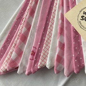 Può includere: Un mucchio di ritagli di stoffa rosa e bianca con diversi motivi, tra cui pois, gingham e disegni floreali. I ritagli di stoffa sono disposti a ventaglio e hanno uno sfondo bianco. Un'etichetta di carta marrone con il testo "De Letscht Schrei" è visibile nell'angolo in alto a destra dell'immagine.