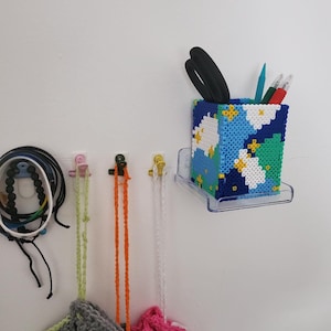 May include: A colourful beaded pencil holder with a blue, green, white and yellow design. The holder is attached to a clear plastic wall mount. There are three crocheted bags hanging below the holder, one grey, one pink and one white.