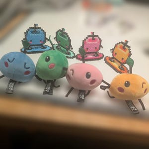 May include: A collection of plush toys and pixelated bead art featuring fruit and drink designs. The plush toys include a blue blueberry, a green apple, a pink strawberry, and an orange peach. The pixel art depicts similar designs in blue, green, pink, and orange.
