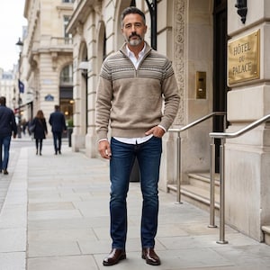 Puede incluir: Un hombre con un suéter beige con un patrón geométrico, una camisa blanca y vaqueros azul oscuro. Está de pie frente a un edificio con un letrero dorado que dice "HOTEL DU PALACE". Lleva botas de cuero marrones.