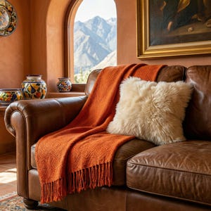 May include: A cozy living room scene featuring a brown leather sofa with an orange throw blanket and a fluffy cream-colored pillow. Decorative pottery and a painting adorn the room, with a mountain view visible through a window.