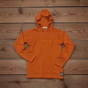 Op de afbeelding: Een okerkleurige trui met lange mouwen en capuchon, voorzien van een fijngebreide textuur. Het kledingstuk heeft een capuchon met een V-vormig halsdetail en een klein, rechthoekig zwart-wit patroonlabel op de rechteronderzoom.