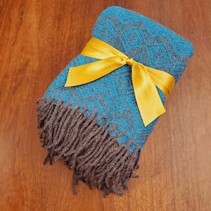 Op de afbeelding: Een turquoise en bruine geweven deken met franjes, gebonden met een geel satijnen lint.