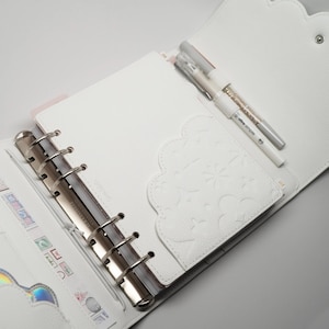 May include: White leather planner with a silver ring binder and a white pocket with a decorative design. Two white pens are inserted in the pocket.