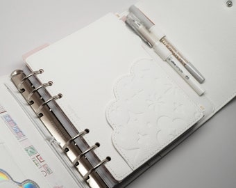 May include: White leather planner with a silver ring binder and a white pocket with a decorative design. Two white pens are inserted in the pocket.