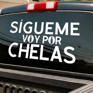 May include: Black truck window with white text that reads "SÍGUEME VOY POR CHELAS". The truck has a red brake light and is parked outside. The text is in a bold, sans-serif font.