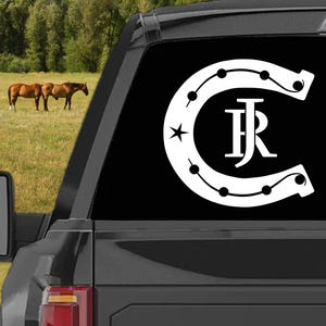 May include: A white horseshoe decal with the letters "JR" inside, a star, and small circles. The decal is on the rear window of a black vehicle. Horses graze in a field in the background.