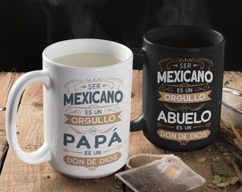 May include: Two white ceramic mugs with black handles. The mugs have black text on them in Spanish. The text on the left mug reads "Ser Mexicano es un Orgullo Papa es un Don de Dios". The text on the right mug reads "Ser Mexicano es un Orgullo Abuelo es un Don de Dios". The mugs are sitting on a wooden surface with a tea bag and other items.