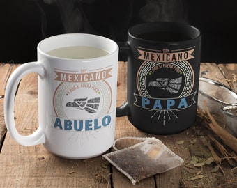 May include: Two white ceramic mugs with black handles. The mugs have a black and white design with the text "Soy Mexicano y por si fuera poco." One mug says "Abuelo" and the other says "Papa". The mugs are on a wooden table with a tea bag and loose tea leaves.