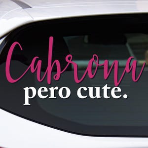 May include: A white car's rear window displays a vinyl decal. The decal features the words "Cabrona" in a cursive, magenta font above the words "pero cute" in a white, sans-serif font.