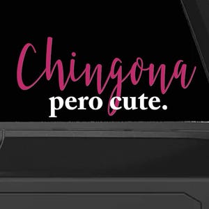 May include: A black vehicle's rear window displays a pink decal with the words "Chingona pero cute." in a cursive font. The phrase "pero cute" is in a smaller, white, sans-serif font.