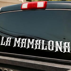May include: Black truck window with a white decal that reads "LA MAMALONA" in a decorative font. The truck bed is visible below the window, with a red brake light above.