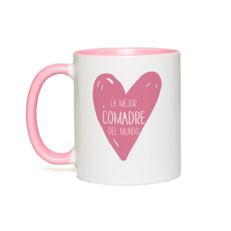 May include: White ceramic mug with a pink rim and handle. The mug features a pink heart with the text "LA MEJOR COMADRE DEL MUNDO" in white.
