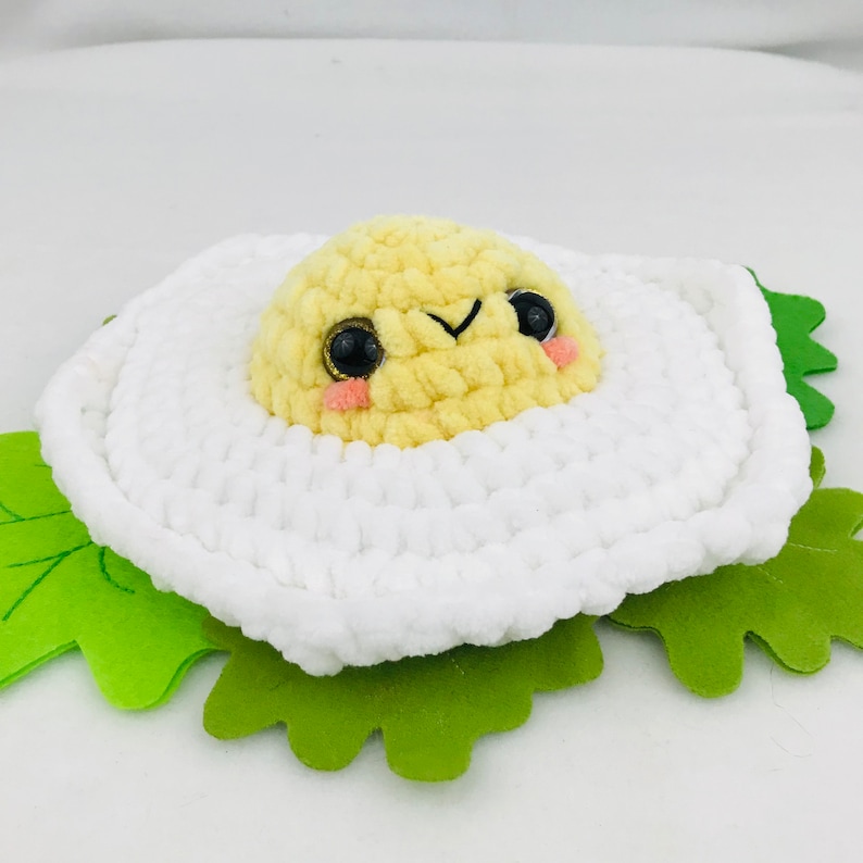Puede incluir: Un juguete de peluche de ganchillo con forma de huevo frito con yema amarilla y clara de huevo blanca. La yema tiene una carita linda con ojos negros y rubor rosado. El huevo est&aacute; sentado sobre una hoja de fieltro verde.