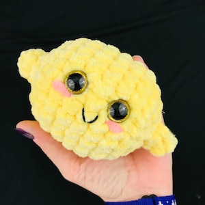 May include: A yellow crocheted lemon plush toy with a smiling face and large black eyes with gold glitter accents.