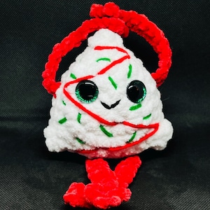 May include: A white plush ornament shaped like a Christmas tree with green and red accents. The ornament has large, glittery eyes and a smiling face. A red loop is attached to the top for hanging, and red plush feet are at the bottom.