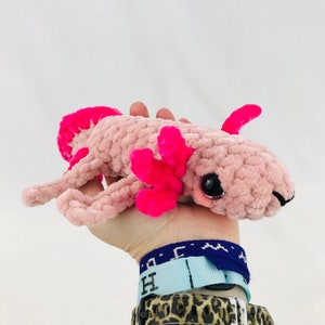 May include: A pink and fuchsia crocheted axolotl plush toy with large black eyes. The axolotl is being held in a hand.