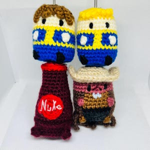 May include: Crocheted figures of two characters with yellow, blue, and brown details, and two other figures: a maroon bottle with "Nuka" text and a figure wearing a cowboy hat. The figures are handmade.